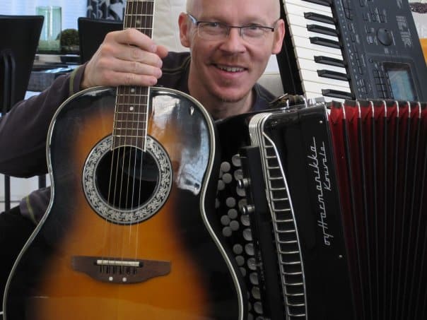 Etualalla kitara, jota Jukka Väisänen pitelee, sylissään harmonikka ja taustalla pystyssä kosketinsoittimet