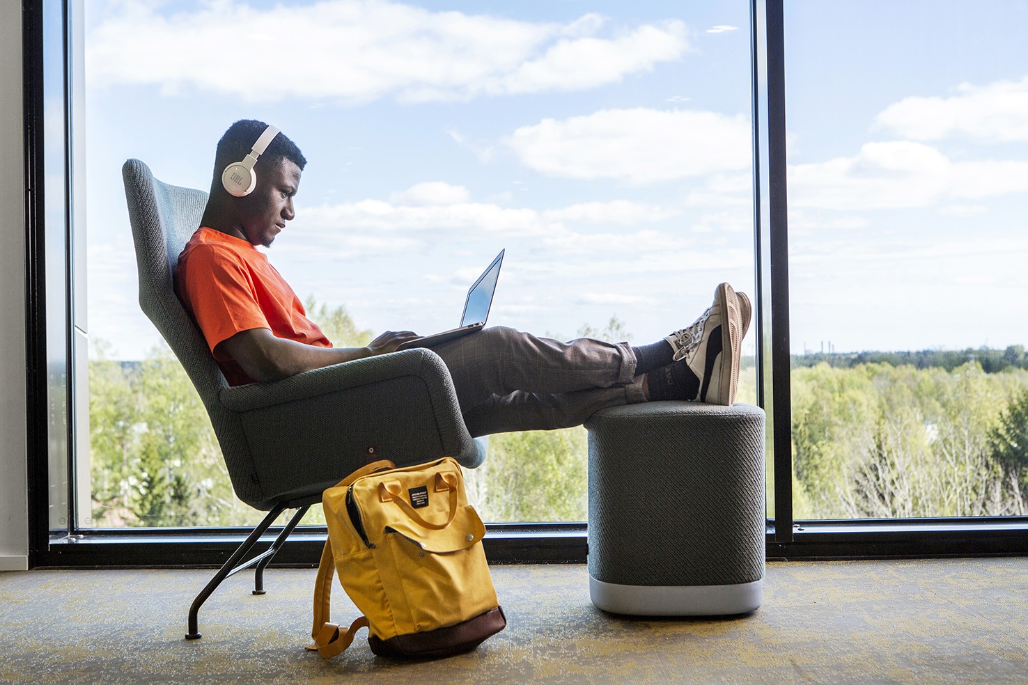 Opiskelija istuu taaksepäin nojaten, jalat rahilla, läppäri sylissä ja kuulokkeet korvilla. Vieressä lojuu keltainen reppu.