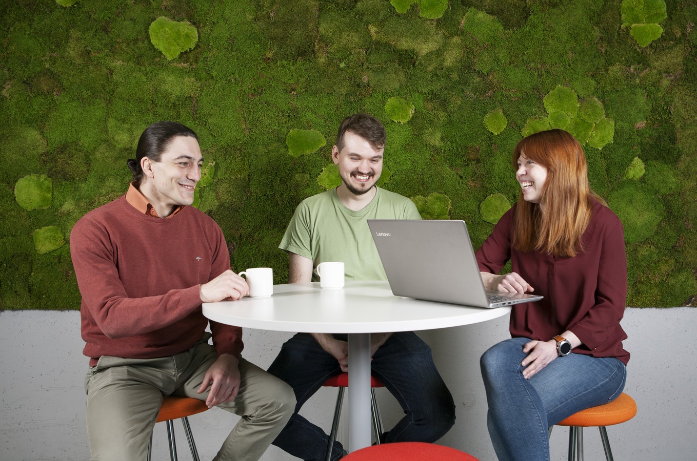 Pyöreän pöydän ympärillä kaksi mies- ja yksi naisoletettu, miehillä kahvikupit edessä pöydällä, naisella läppäri. Taustalla sammalesta tehty viherseinä.