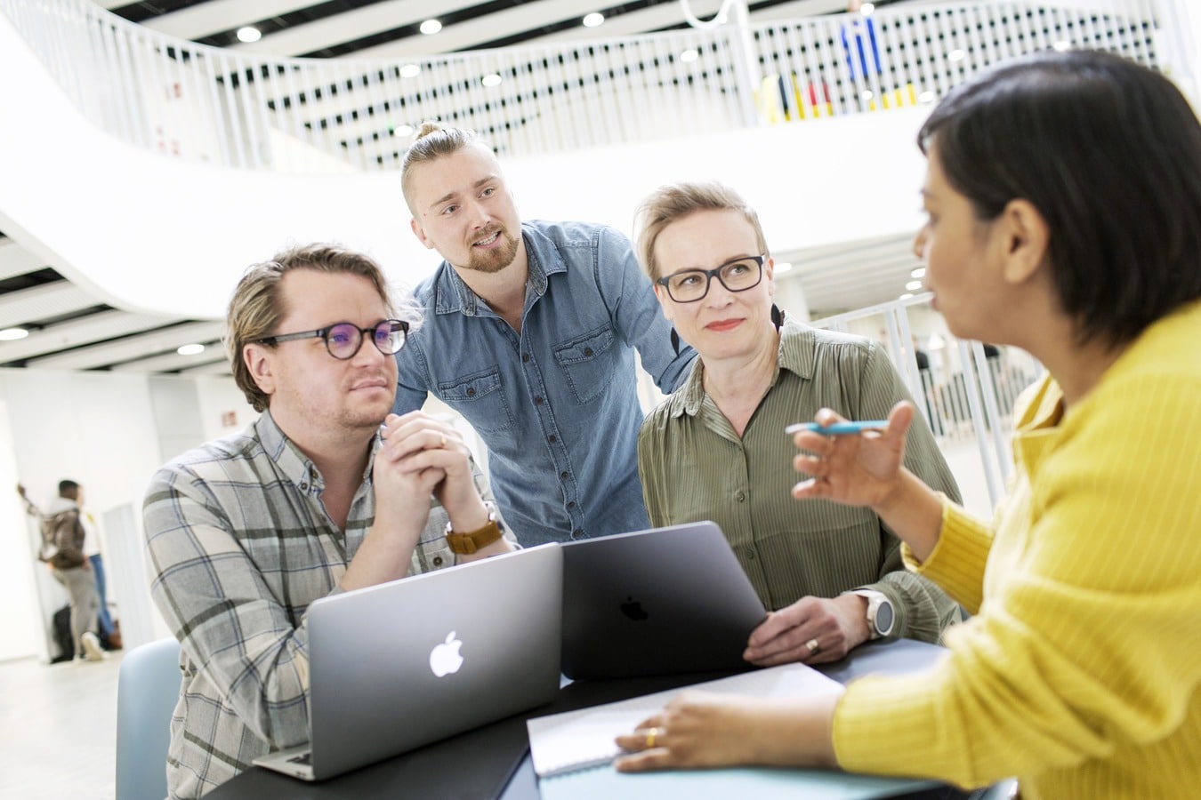 Kaksi henkilöä istuu läppärit edessään, heidän takana seisoo yksi henkilö kumartuneena kohti puolittain etualalla näkyvää neljättä henkilöä kohti.