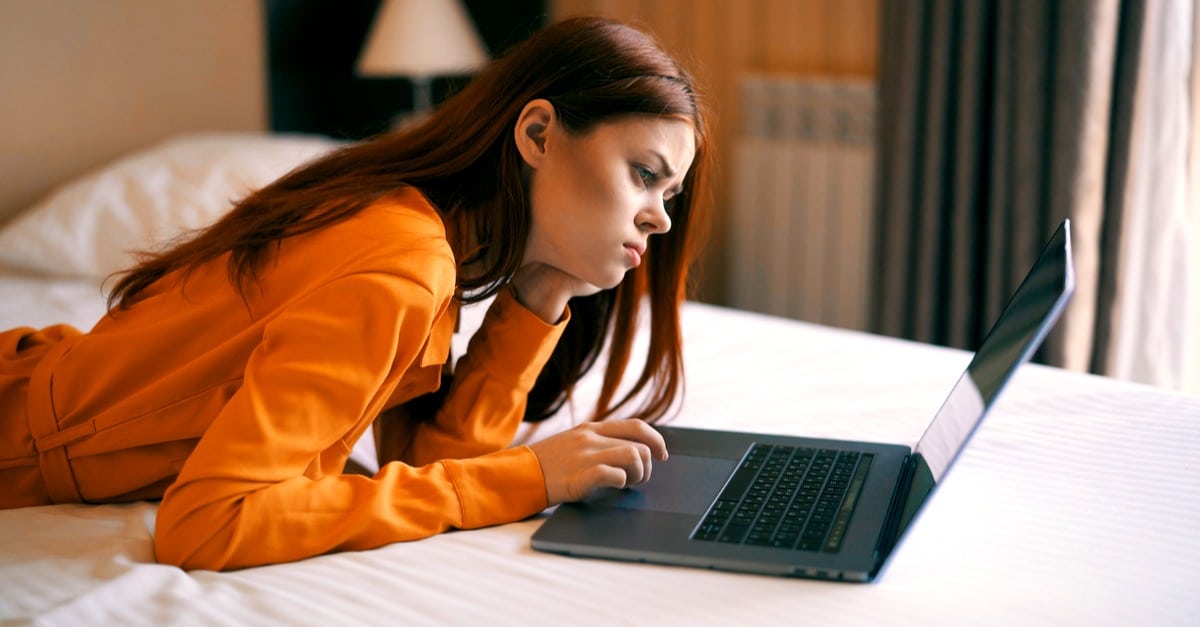 Makuuhuoneessa oranssipaitainen nainen makaa mahallaan sängyllä läppärin äärellä
