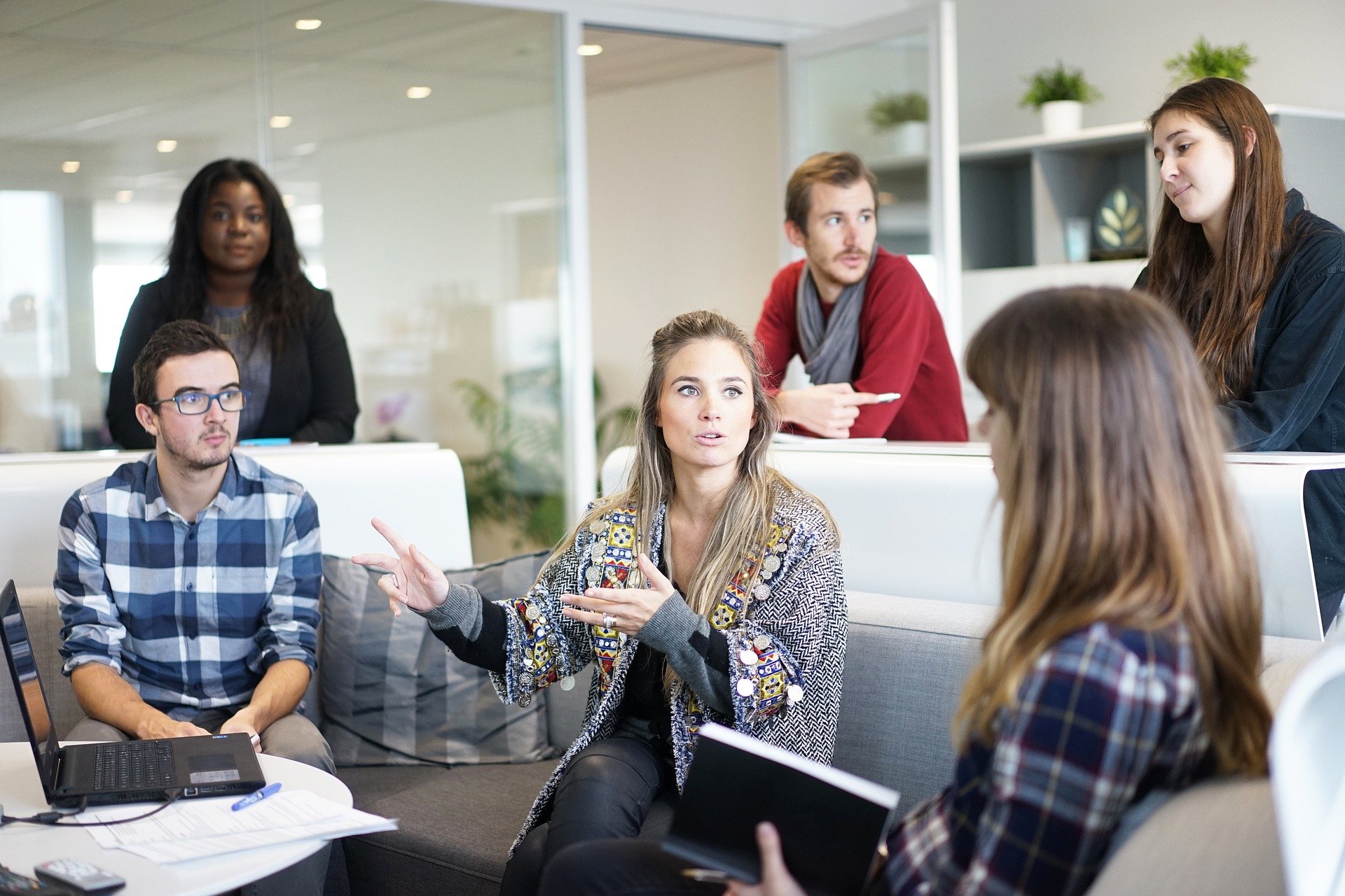 Young adults at office, three seated around a table and three standing behind them, discussing with each other.