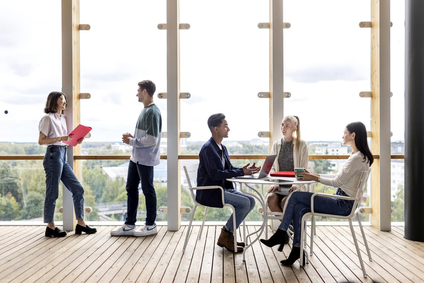 Viisi opiskelijaa Myllypuron kampuksen lasitetulla terassilla, kolme istumassa pöydän äärellä ja keksi seisomassa vastakkain ikkunan äärellä.