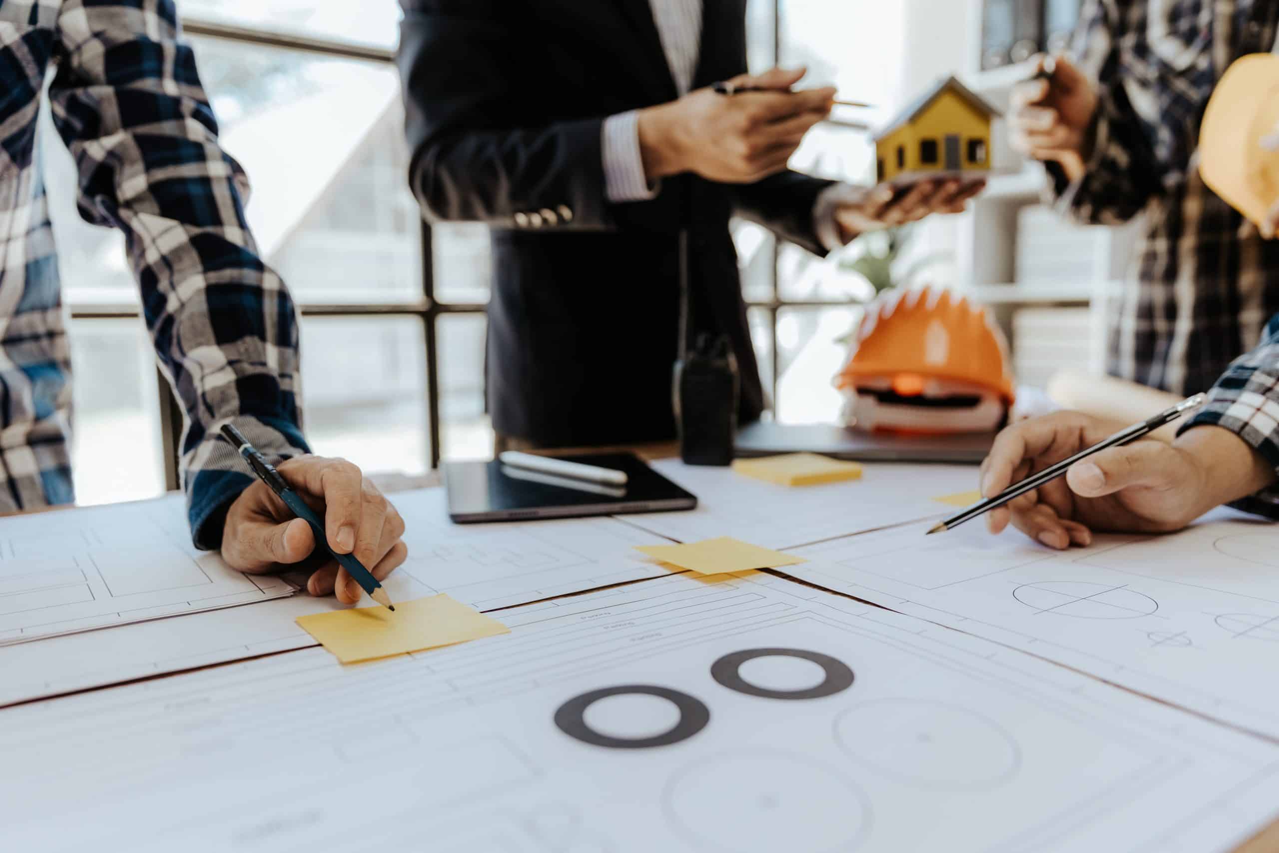 Rakennuspiirrustuksia ja suojakypärä pöydällä, jonka äärellä kolme ihmishahmoa, näkyvillä käsiä ja keskivartaloa.