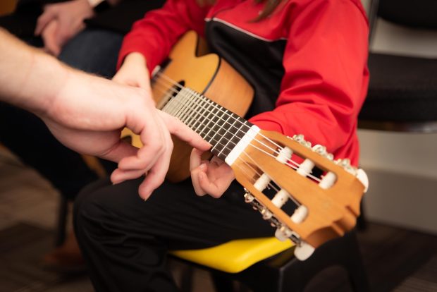 Kuvassa lapsi soittamassa kitaraa ryhmässä, ohjaaja neuvoo E-kielen näppäämisessä.