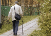 Vanha mieshenkilö kävelee kepin kanssa ulkona poispäin kuvaajasta.
