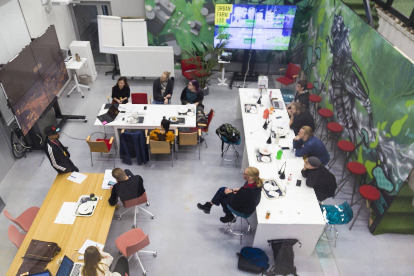 Ylhäältä kuvattu ihmisiä istumassa pöytien äärellä, yksi seisomassa vasemmalla, taustalla diginäyttöjä ja Metropolian Myyrmäen kampuksen Urban Farm Lab -tilan kasveja.