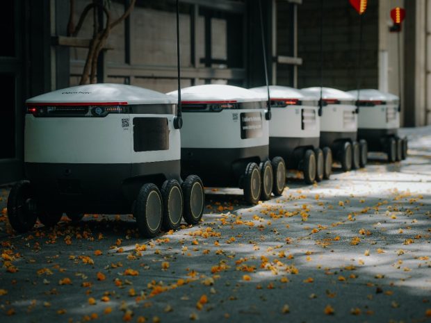 Viisi autonomista robottia liikkuvat jonossa kadulla.