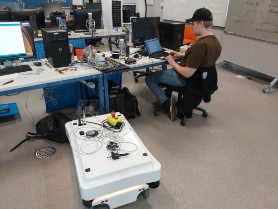 Henkilö istuu automaatiolaboratoriossa tietokoneen ääressä ja lattialla vieressä on robotti.