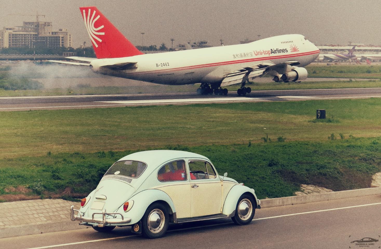 Etualalla pieni auto ajaa tiellä. Takana lentokone etenee kiitoradalla samaan suuntaan auton kanssa.