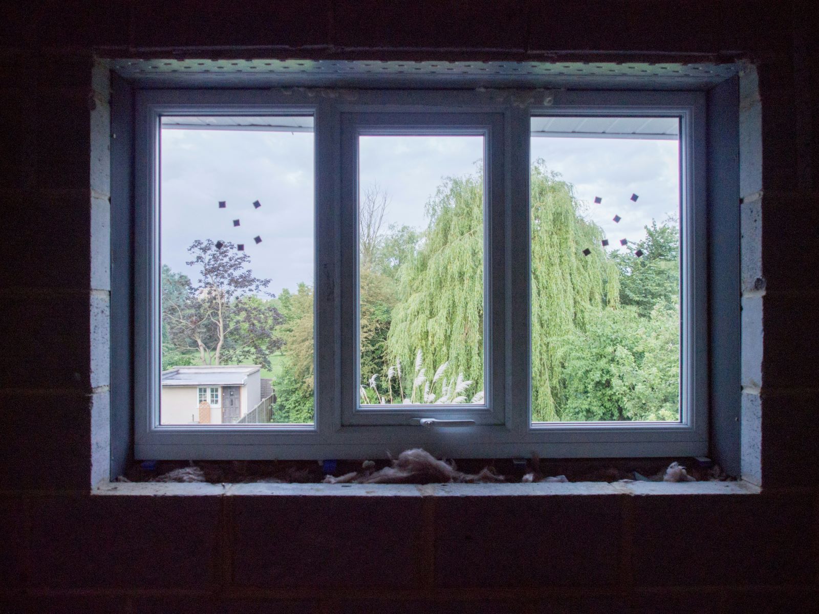 Sisältä ulospäin kuvattu kolmiruutuinen ikkuna, jonka eristeet ovat näkyvillä. Taustalla vihreitä lehtipuita ja talo.