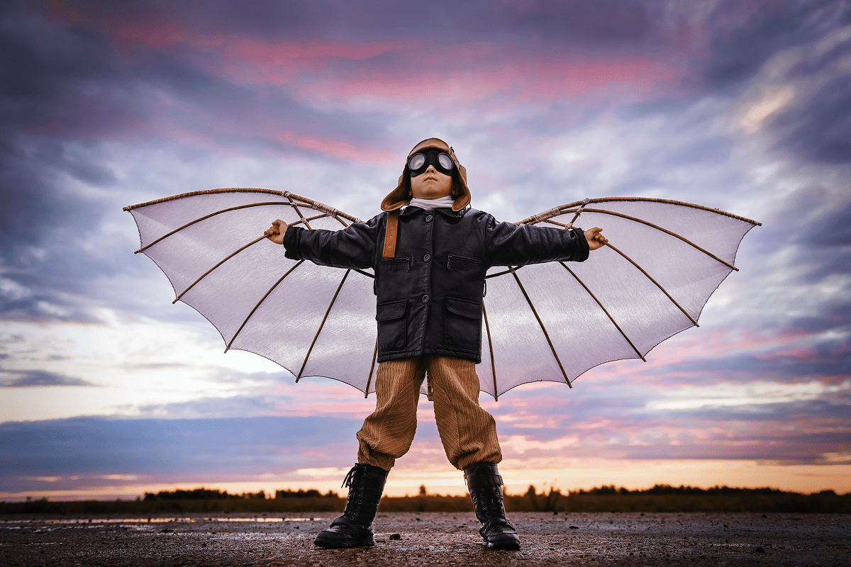 Lapsi seisoo haara-asennossa ulkona, tekosiivet selässä ja kypärä sekä lentäjänlasit päässä, katse kohti taivasta, taustalla peltoa ja pilvinen taivas
