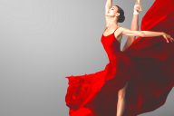 Ballerina seisoo yhdellä jalalla sivuprofiilissa kärkitossuilla punaisessa hulmuavassa mekossa, kädet avoinna katse ylöspäin