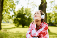 Nuori hymyilevä naishenkilö istuu viheröivässä puistossa silmät suljettuna ja kuulokkeet päässä