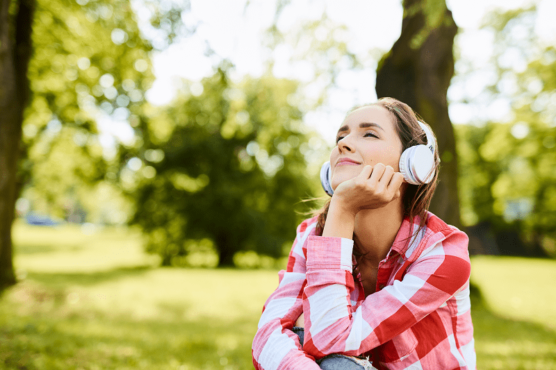 Nuori hymyilevä naishenkilö istuu viheröivässä puistossa silmät suljettuna ja kuulokkeet päässä