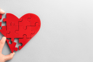 Heart-shaped red puzzle on plain grey surface, two hands holding pieces