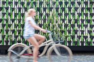 Taustalle kameran tarkentaman monien pienten istutusruukkujen muodostama viherseinä ulkotiloissa, sen edustalla sumeana naishenkilö pyöräilemässä vaalealla perinteisellä polkupyörällä
