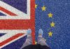 Puolikas Iso-Britannian lippu ja puolikas EU:n tähtilippu maalattuna maahan, henkilö seisoo keskellä, kuvattu ylhäältä niin että vain jalat ja kengät näkyvät.