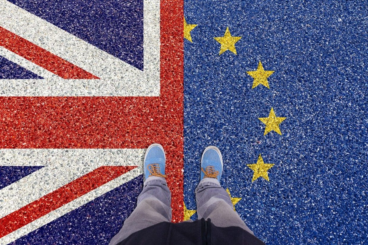 Puolikas Iso-Britannian lippu ja puolikas EU:n tähtilippu maalattuna maahan, henkilö seisoo keskellä, kuvattu ylhäältä niin että vain jalat ja kengät näkyvät.