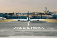 Lentokone takaapäin, kiitoradan etualalla teksti Helsinki, horisonttiin taustakuvaksi liitetty Helsingin keskustan siluetti mereltä päin kuvattuna, tuomiokirkon torni oikealla sivulla esillä.