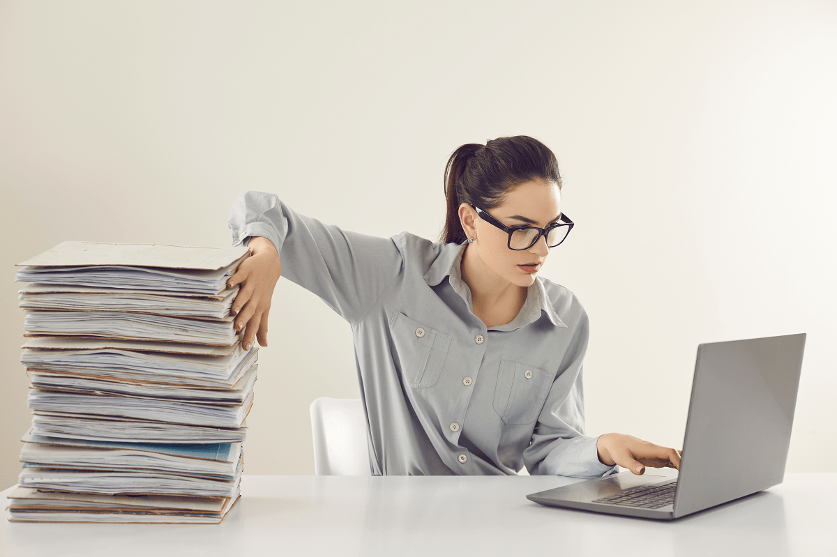 Työpöydän takana istuva nainen pitelee korkeaa paperipinoa pystyssä toisella kädellä ja on kohdistanut katseensa edessään olevaan kannettavaan tietokoneeseen.