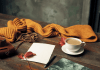 Kuvassa syksyn paletissa pöydällä sinapinkeltainen kaulahuivi, muistikirja, punaiset lehdet, kahvijuoma valkoisessa posliinimukissa tassin päällä, kameran nahkakotelo