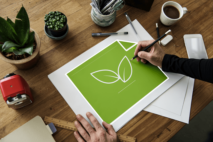 Työpöydällä kyniä, pieniä kasveja, kahvikuppi ja keskellä vihreä paperi, jonka molemmin puolin näkyvät kädet, oikea pitelee kynää.