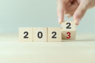 Neljässä pöydälle asetetussa puupalikassa etualalla numeroista muodostuva vuosiluku 2022, viimeistä palikkaa pitelevät sormet paljastavat numeron olevan vaihtumassa kakkosesta kolmoseksi 2023
