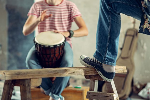 In front: a foot in street shoe rests against railing, a male torso is seated and drumming.