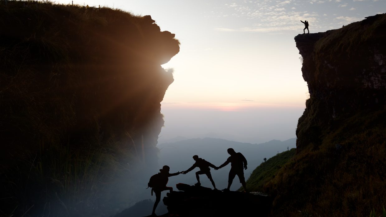 Three hikers pulling each other upwards on a mountain top in the front, one person standing alone on top in the background, silhouette of mountain and misty skies in the back.