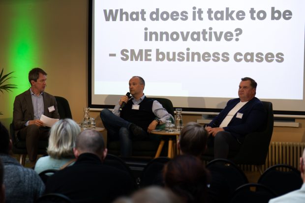 Peter Nyman haastattelee Erno Launoa ja Mikko Karellia. Miehet istuvat rinnakkain tuoleilla yleisön edessä ja heidän takanaan on valkokankaalle heijastettu teksti What does it take to be innovative - SME business cases.