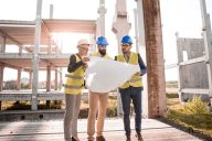 Kolme henkilöä seisoo rakennustyömaamiljöössä kypärät päässään ja tarkastelevat rakennuspiirustuksia, joita keskimmäinen henkilö pitää käsissään edessään.