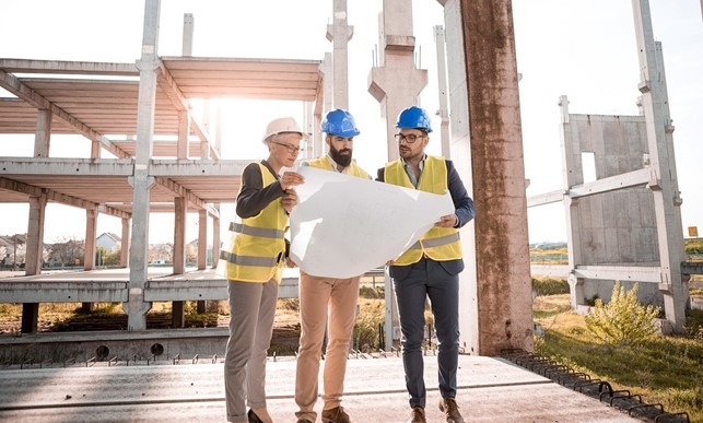 Kolme henkilöä seisoo rakennustyömaamiljöössä kypärät päässään ja tarkastelevat rakennuspiirustuksia, joita keskimmäinen henkilö pitää käsissään edessään.