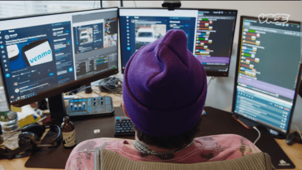 Violettipipoinen henkilö istuu selin kameraan ja tuijottaa kolmea tietokoneruutua, jotka on hänen edessään.