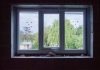 Sisältä ulospäin kuvattu kolmiruutuinen ikkuna, jonka eristeet ovat näkyvillä. Taustalla vihreitä lehtipuita ja talo.