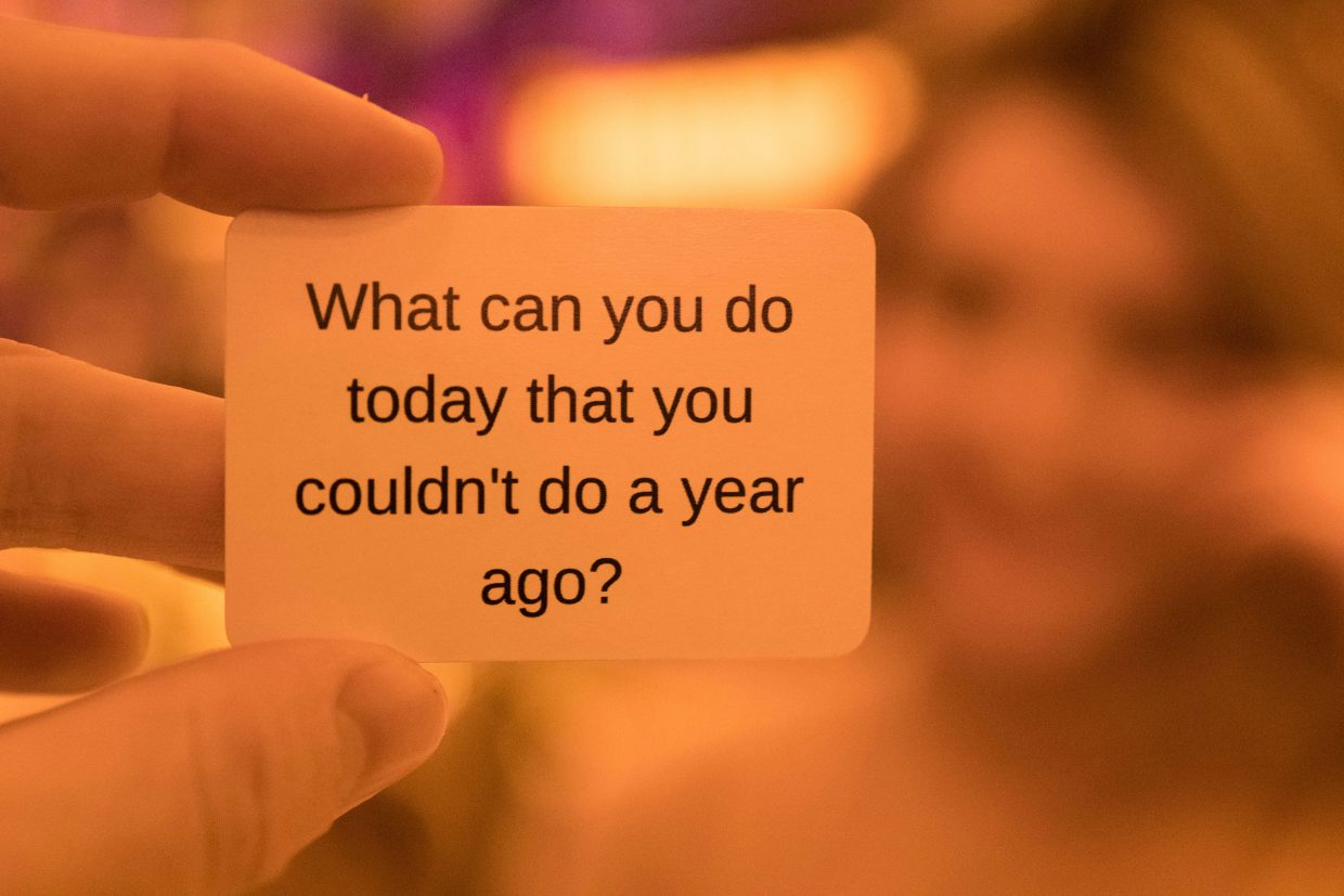 In background a blurred human face, in center closeup of fingers holding a card reading What can you do today you couldn't do a year ago? with strong sepia orange hue.