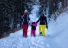 Luminen metsämaisema, jonka keskellä lumen peittämää polkua pitkin kävelee reunoilla kaksi aikuista, pidellen välissään kävelevää lasta käsistä.