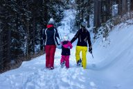 Luminen metsämaisema, jonka keskellä lumen peittämää polkua pitkin kävelee reunoilla kaksi aikuista, pidellen välissään kävelevää lasta käsistä.