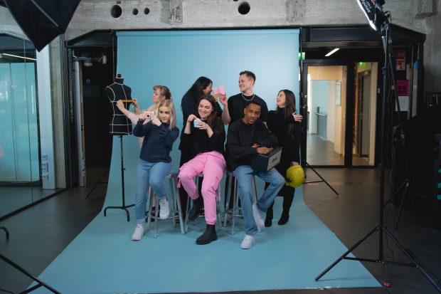 Ryhmävalokuva, taustalla vaaleansininen kangas, näkyvillä valaisimen jalusta, 7 nuorta aikuishenkilöä poseeraa arkivaatteissa seisten ja korkeilla tuoleilla istuen.