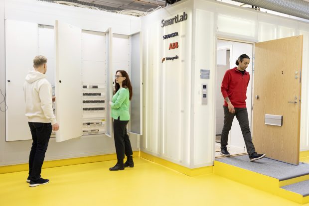 Two people standing in opposite corners, shorter in green and taller in red shirt, of room, yellow floor and white walls with SmartLab texts