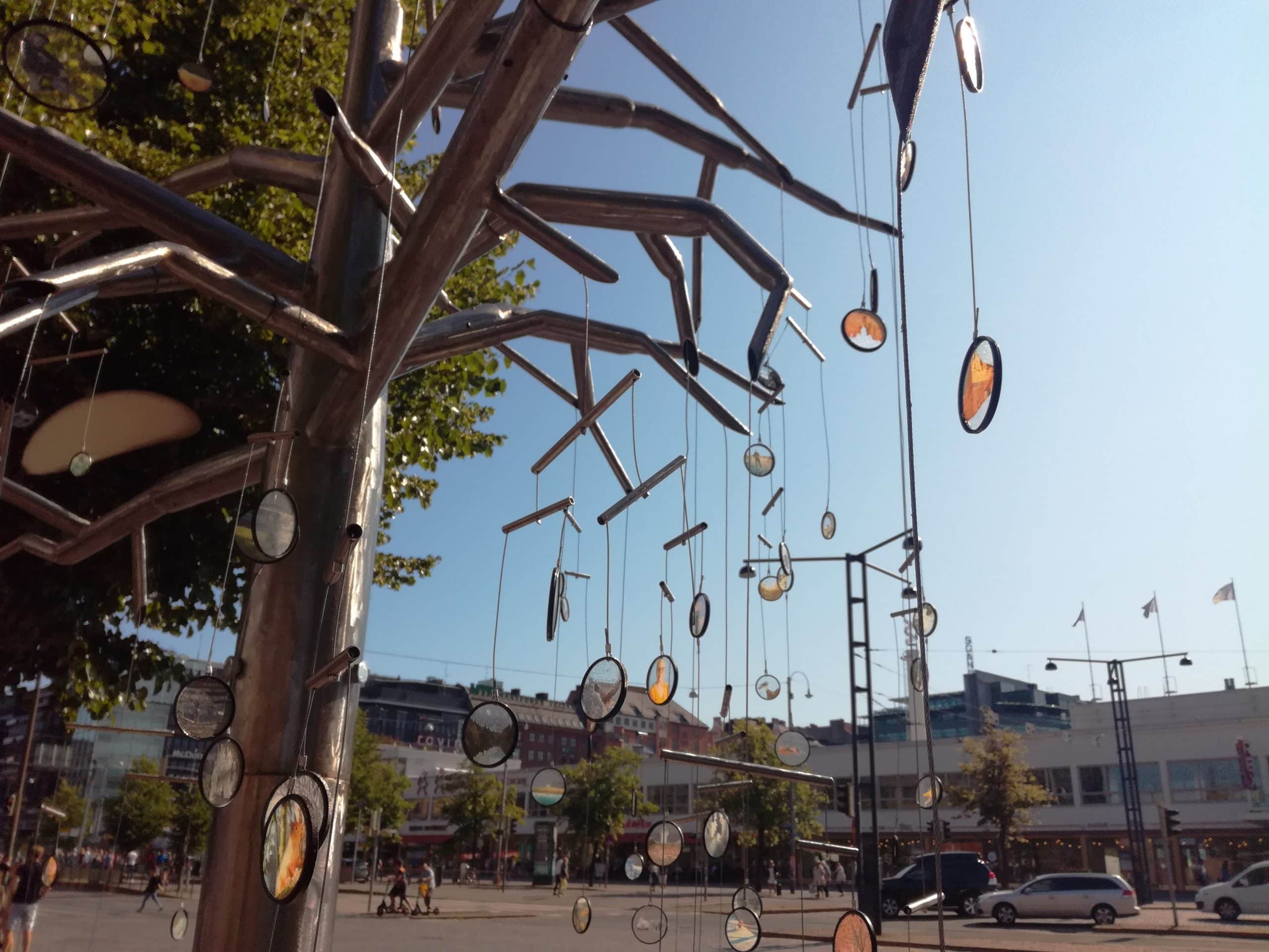 http://A%20summer%20street%20view%20from%20the%20ground%20overlooking%20Mannerheimintie%20street%20in%20Helsinki%20and%20the%20Lasipalatsi%20building%20and%20other%20houses%20on%20the%20other%20side%20of%20the%20street.%20on%20the%20foreground%20is%20a%20tree-like%20art%20installation%20with%20many%20translucent%20circular%20lenses%20with%20different%20photos%20or%20images%20hanging%20from%20the%20branches.%20Three%20cars%20are%20seen%20on%20the%20picture%20as%20well%20as%20several%20pedestrians%20and%20two%20people%20with%20electric%20kickboard%20crossing%20the%20street.