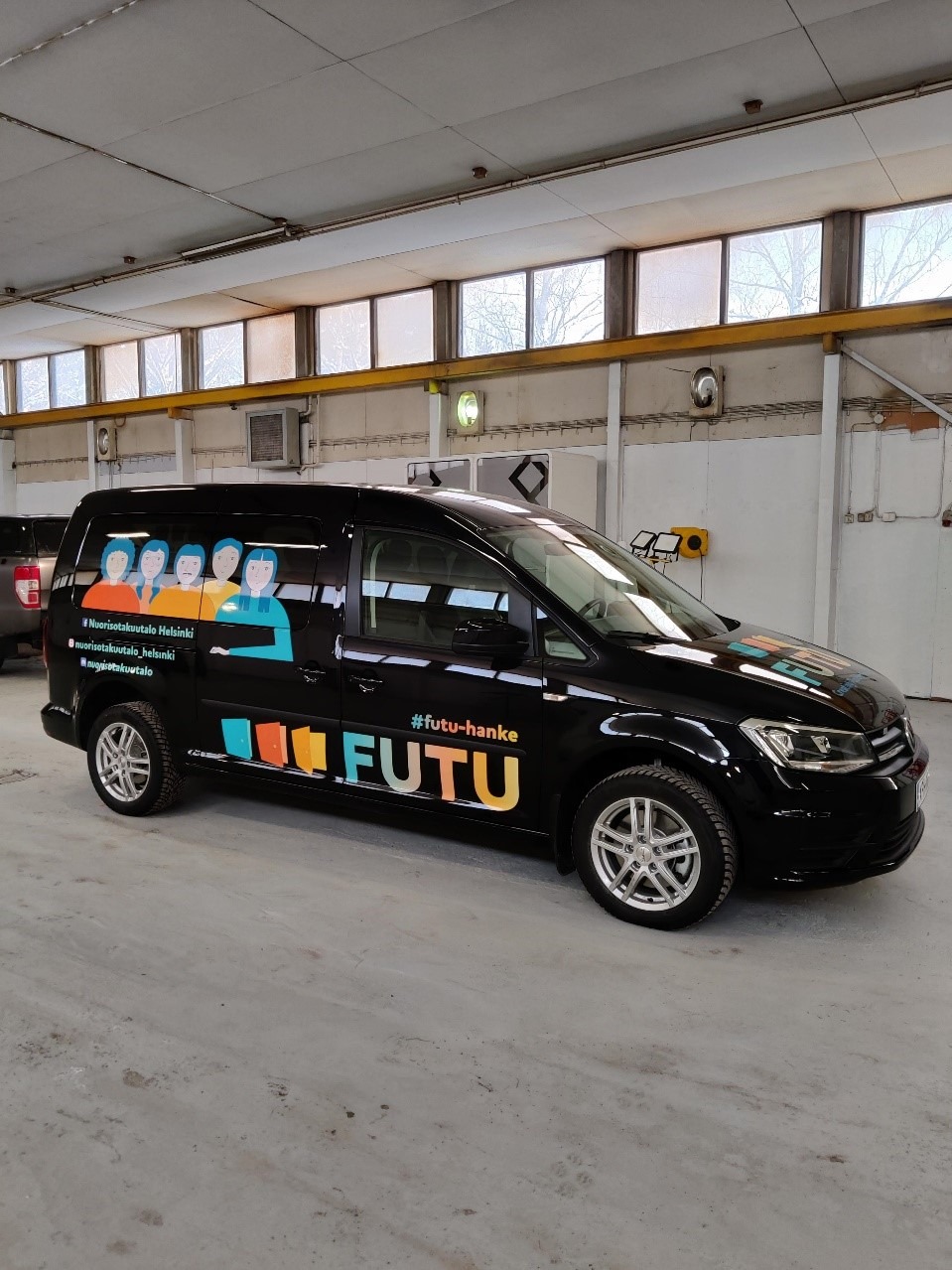 Autossa sivusta kuvattu tumma iso tila-auto, jossa FUTU-hankkeen kirjavat tarrat kyljissä.