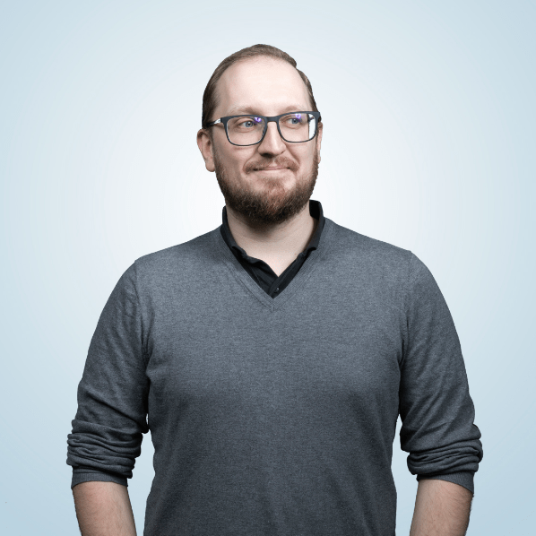 Silmälasipäinen parrakas mieshenkilö puolikuvassa vaaleaa taustaa vasten tummanharmaassa paidassa.