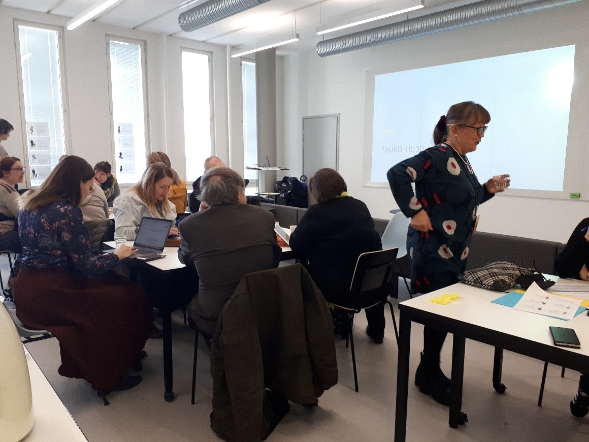 Ryhmä ihmisiä luokkahuoneessa pöydän ääressä, opettaja seisomassa.