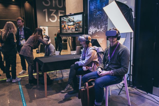 Kaksi henkilöä kokeilemassa VR-sisältöjä Match Up 2019 -tapahtumassa. He istuvat tuoleilla VR-lasit päässään. Alue on rajattu lattiateipillä. Taaempana näkyy kolmas henkilö VR-lasit päässä.