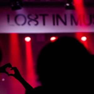Kuvassa Lost In Music- banderolli keikkayleisön keskeltä, kameran tarkennus osuu osallistujan käsien muodostamaan sydämeen.