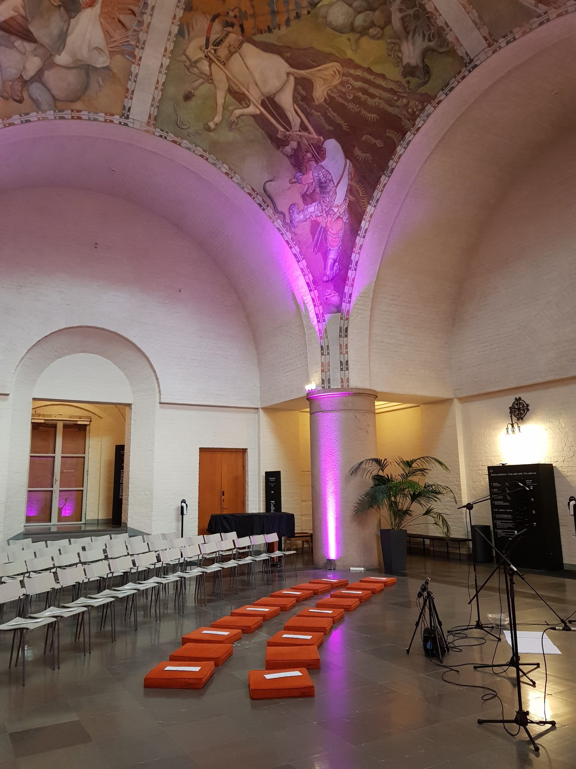 Kuva on Kasnallismuseon keskihallista. Kuvassa näkyy hyvin koristeelliset kattomaalaukset. Lattialle on asetettu tuoleja riviin ja niiden eteen oransseja istuintyynyjä. Niiden edessä on mikkitelineitä.