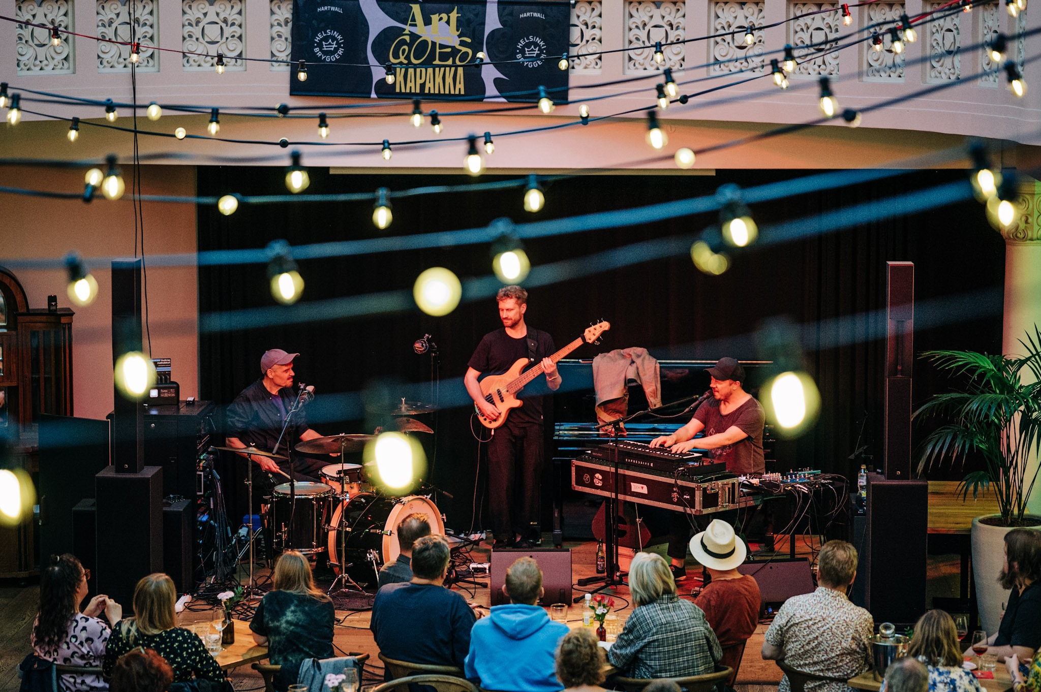 Art Goes Kapakka keikka Sofia Helsingissä. Kuvassa lava, jossa soittaa artistin Tuomo yhtye: pianisti, kitaristi ja rumpali. Kuvan etualalla istuu yleisöä. 