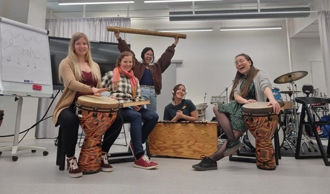 Viisi innokasta opiskelijaa, neljä reunimmaista istumassa ja yksi keskellä seisomassa luokkahuoneessa.