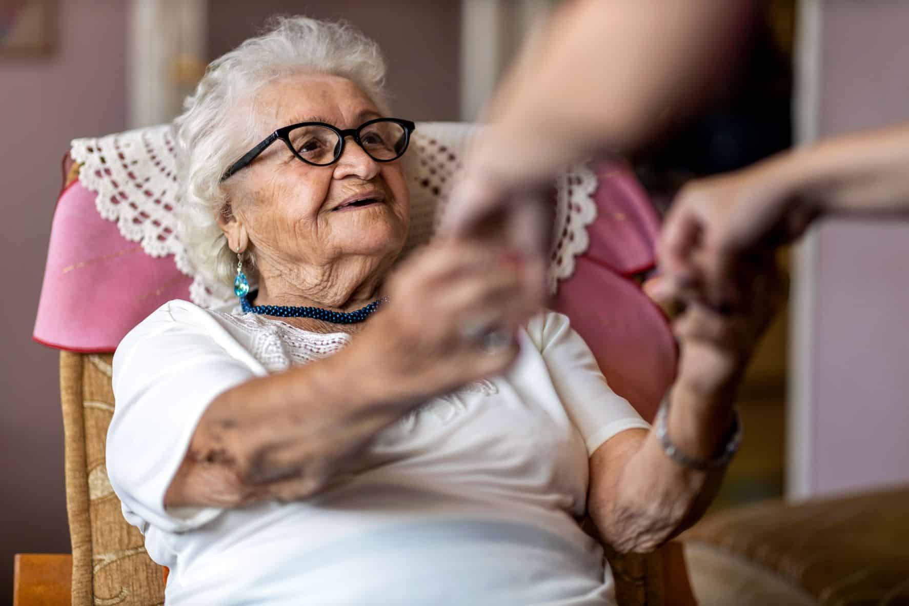 Iäkäs silmälasipäinen ja harmaakiharainen naishenkilö istuu valkoinen T-paita päällään nojatuolilla kädet ojennettuna eteenpäin, toisen ihmisen kädet tarttumassa vanhuksen käsiin.